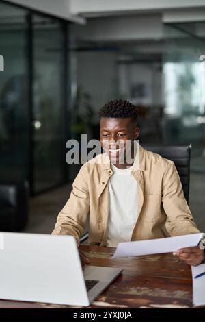 Happy young African business man holding documents using laptop at work. Stock Photo