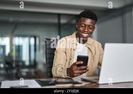 Happy busy African business man using phone working on laptop at office desk. Stock Photo
