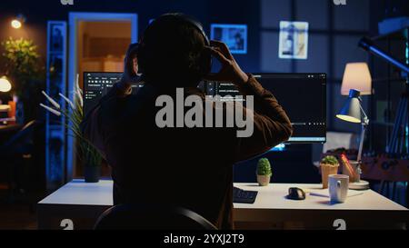 Technician putting headphones on while using coding to fortify firewalls, teleworking for IT company. Computer scientist listening music and upgrading binary code scripts in home office, camera B Stock Photo