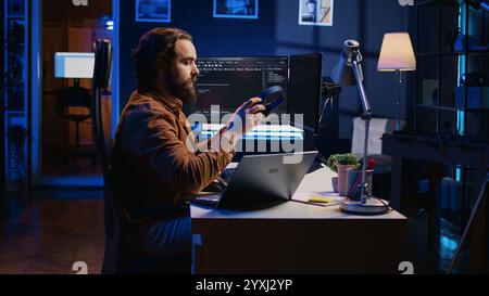 Developer writing lines of code on multimonitor PC setup during remote job shift. IT expert doing multitasking in neon lit home office, listening music, drinking coffee, camera B Stock Photo