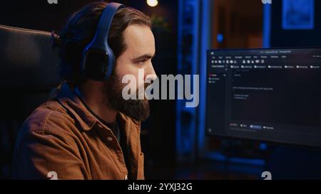 Computer scientist listening music while writing lines of code during remote job shift in home office. IT specialist enjoying audio podcast, coding on desktop PC, camera B close up Stock Photo