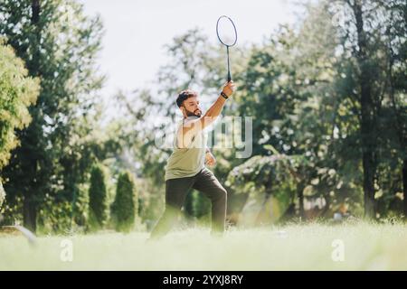 Young man enjoys playing badminton in a grassy park. The setting is sunny with lush green trees, capturing an active outdoor lifestyle. Stock Photo