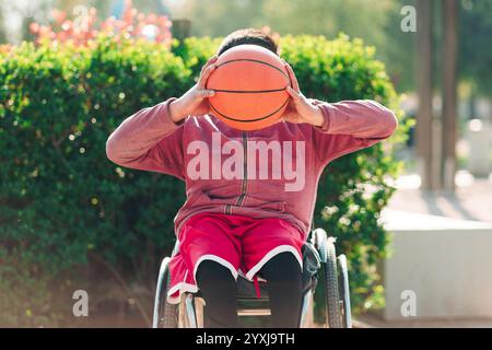Young Latino athlete in wheelchair, posing, holding a basketball and hiding behind, in a park in the city outdoors Stock Photo