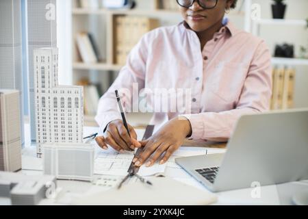 African woman architect focused on drawing city blueprint with scale models. Professional workspace includes laptop and drafting tools, highlighting creativity and precision in architectural design. Stock Photo