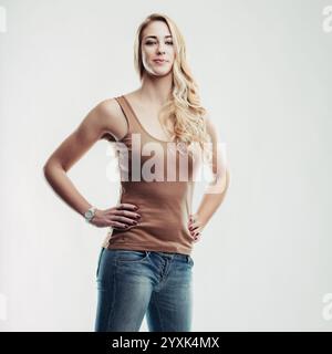 Young woman with long blond hair, wearing a sleeveless top and jeans, stands confidently with her hands on her hips, exuding strength and self-assuran Stock Photo