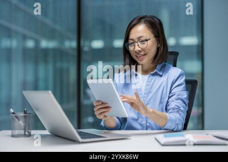 Asian woman dressed professionally, using a tablet at her desk in office. She appears focused and engaged, demonstrating concepts of technology usage and productivity in professional environments. Stock Photo