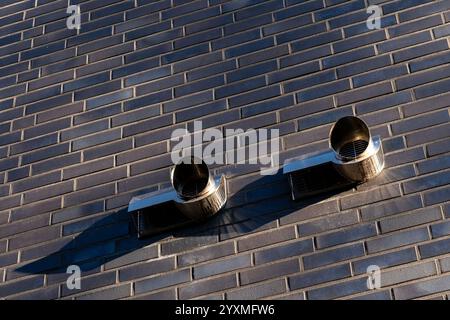 Two metallic ventilation pipes are mounted on a textured brick wall, casting shadows as the sunlight reflects off their surfaces during the afternoon. Stock Photo
