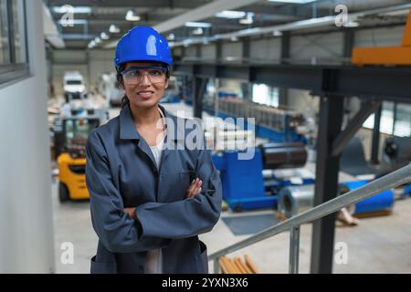 Confident young engineer, an Indian woman, in a blue helmet and gray work coat at a factory, portrait shot. Stock Photo