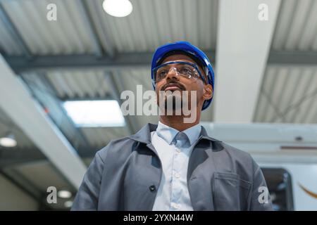 African American engineer, wearing a gray work coat and a blue helmet, controlling production and programming a machine in a factory. Stock Photo