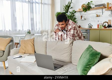 Young man concentrated on his work at a laptop while leaning over a cozy sofa with pillows. Stock Photo