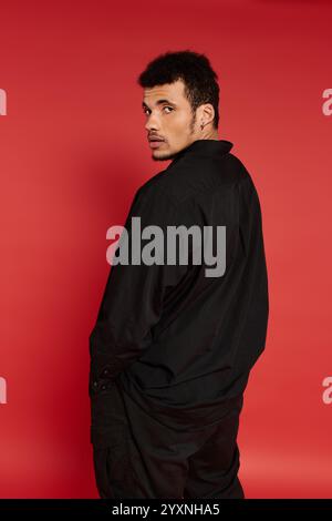 The young man with a stylish look stands confidently against a bright red backdrop. Stock Photo