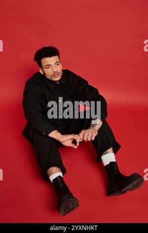 A young man with striking features sits casually on the floor, dressed in a black outfit. Stock Photo