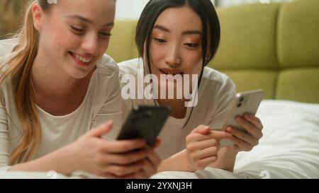Two happy smiling multiracial friends multiethnic women diversity girls female Caucasian Asian Japanese Chinese relaxing on bed together using mobile Stock Photo