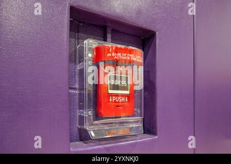 Red fire alarm system pull station under a protective clear plastic cover.  Fire alarm in a gymnasium mounted on wall with purple protective wall pads. Stock Photo