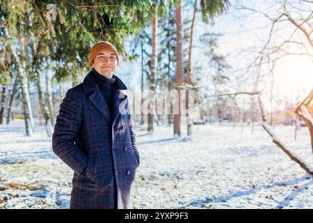 Portrait of young stylish man posing in winter park on sunny day. Guy wearing warm coat enjoying snowy landscape Stock Photo