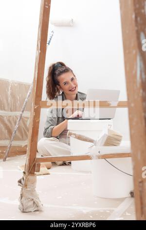 Happy woman using her laptop computer to search for and buy wall paint during her home renovation. She is sitting near a paint bucket and a wooden lad Stock Photo