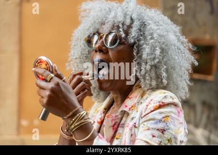 Expressive, black lady with hoary afro hair and sunglasses putting on blue lipstick in sunny city center. Concept: pro-aging, self-confidence Stock Photo