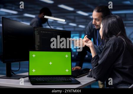 Multiracial team reconfiguring server farm to optimize it for data processing using green screen laptop. Diverse coworkers ensuring data center equipment operates efficiently using mockup notebook Stock Photo