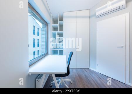 Bright and spacious home office featuring a white desk and chair, large window with outdoor view, and minimalist storage units, AC on the Wall. Modern Stock Photo