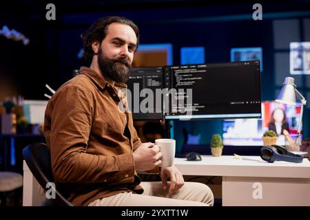 Portrait of cheerful computer engineer enjoying hot beverage while implementing security measures to protect company systems and data. Smiling IT worker drinking coffee and debugging code Stock Photo