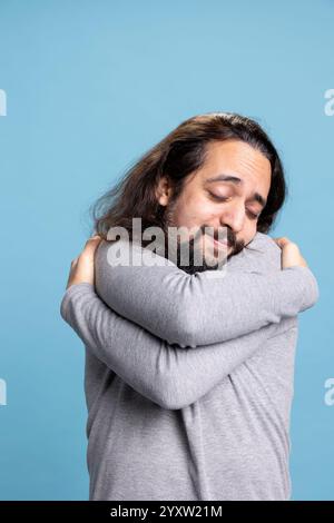 Happy person feeling fulfilled and embracing himself as a form of self love, showing self appreciation and a powerful personality against blue background. Young adult relaxed and confident. Stock Photo