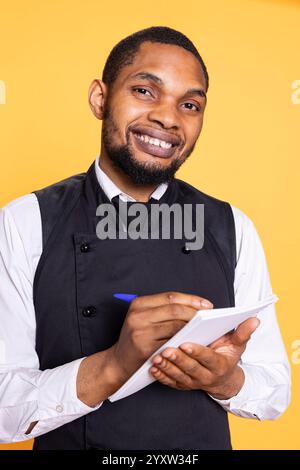 Friendly restaurant waiter taking notes of meal orders for clients, using information on the notepad for the bill. Butler serving people for customer satisfaction against yellow background. Stock Photo