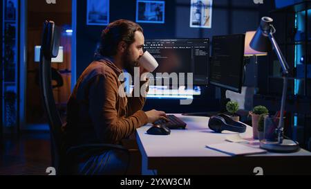 Focused programmer drinking coffee in apartment while solving database errors on computer. Remotely working IT admin concentrated on finalizing coding project in home office, camera B Stock Photo