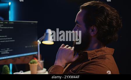 Happy self employed developer in home office doing coding on computer at desk. Joyful IT specialist doing contracted job from apartment, writing lines of code at workstation, camera B close up Stock Photo
