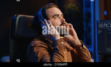 Cheerful programmer listening music while doing web development during remote job shift in neon lit home office, finishing coding project. IT expert enjoying audio podcast, working on PC, camera A Stock Photo