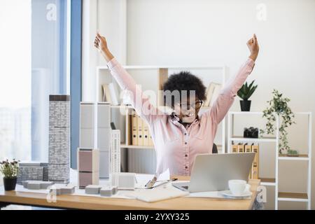 Happy African woman architect celebrates successful city model design in modern office. She exudes joy and accomplishment while working on cutting-edge project with architectural models. Stock Photo