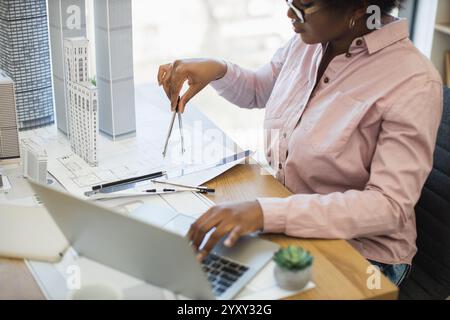 African woman architect using compass to measure dimensions on modern city project model. She is focused on design details at her workstation, surrounded by architecture tools and laptop. Stock Photo