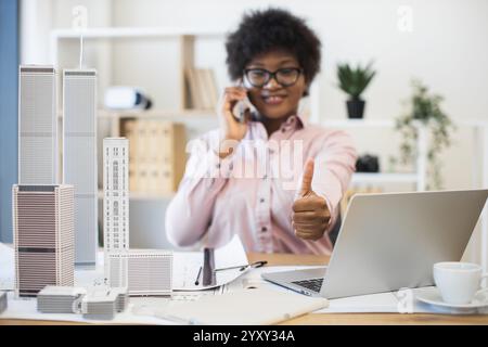 Confident African businesswoman architect having phone conversation about city project. Smiling and giving thumbs up while working with laptop and architectural models in office. Stock Photo