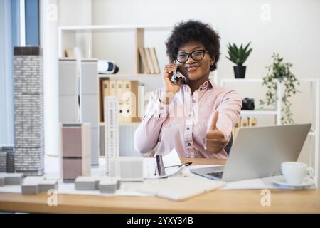 Confident African businesswoman architect having phone conversation about city project. Smiling and giving thumbs up while working with laptop and architectural models in office. Stock Photo