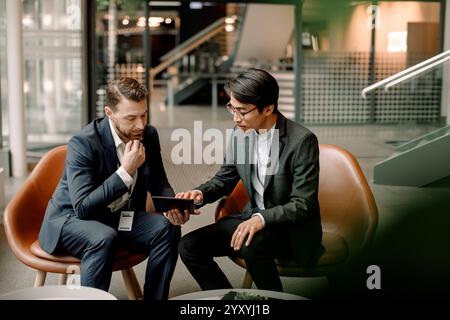Male entrepreneurs discussing over digital tablet while sitting on chairs at convention center Stock Photo