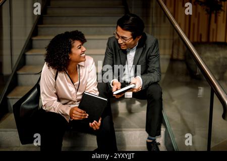Happy male and female business professionals laughing while sitting on staircase at convention center Stock Photo