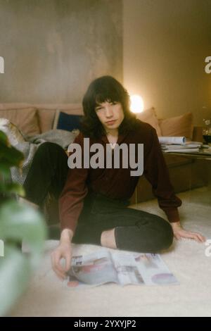 Portrait of young man with long hair sitting on floor near magazine while posing at home Stock Photo