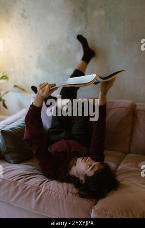 Comfortable young man reading magazine while lying down on sofa at home Stock Photo