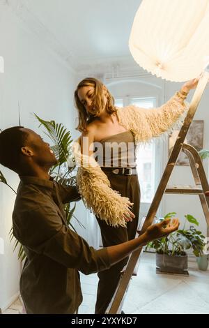 Smiling young man talking with girlfriend standing on ladder at apartment during relocation Stock Photo