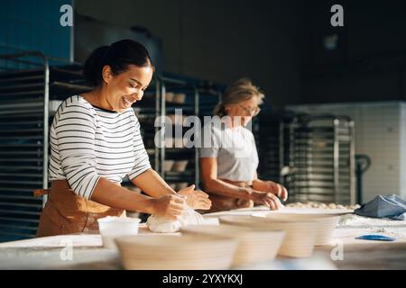 Smiling female baker kneading dough on table while working with colleague at bakery Stock Photo