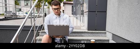 Man is sitting on stairs using laptop, focused on work while enjoying coffee nearby. Stock Photo