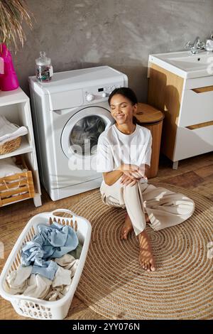 In a modern apartment, a young african american woman relaxes while doing laundry, smiling. Stock Photo