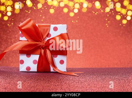 White gift box with polka dots with red bow stands on shiny background. Defocused lights. Christmas, New Year, Saint Valentine, March 8, Mother's Day, Stock Photo