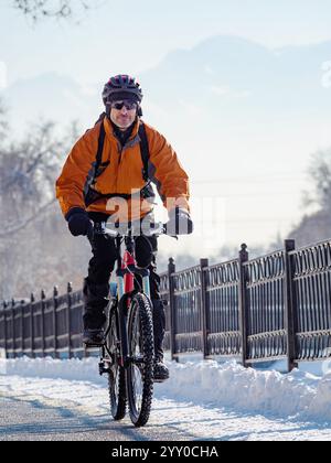 A cyclist riding through a snowy city on a sunny day. You can see the mountains in the background Stock Photo