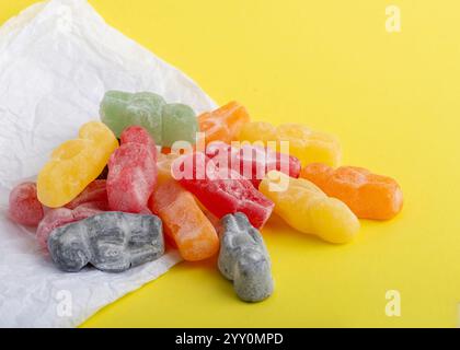 pile of vibrant jelly baby candies in red, green, orange, yellow, and black rests on crinkled white paper against a bright yellow background, Stock Photo