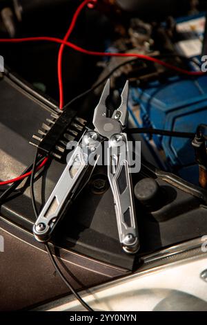 A multitool with pliers is placed on a car engine, surrounded by red and black wires, suggesting a repair or maintenance task. Stock Photo
