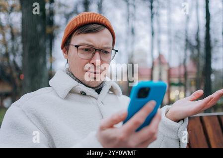 Stylish man wearing white jacket and orange beanie looking confused while reading surprising news on his blue smartphone, sitting on a bench in a park Stock Photo