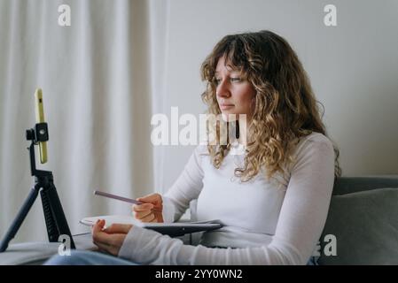 Curly haired female teacher recording online course using smartphone on tripod, holding pencil and notebook while sitting on sofa at home Stock Photo