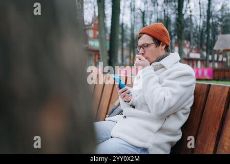 Young man wearing a white jacket and orange beanie sits on a park bench, using his smartphone while coughing and appearing unwell Stock Photo