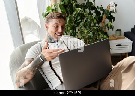 A young man with tattoos smiles and gestures while engaged in a video call in his cozy living room. Stock Photo