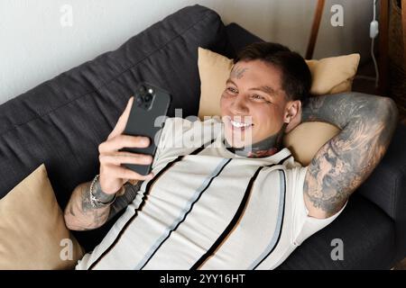 Handsome young man with tattoos smiles while enjoying his time on the phone at home. Stock Photo